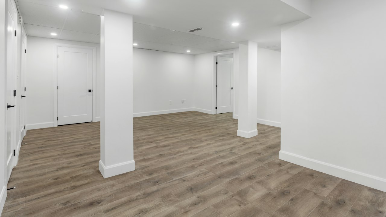 An empty finished basement room with white walls and wood-look flooring, unlit and quiet